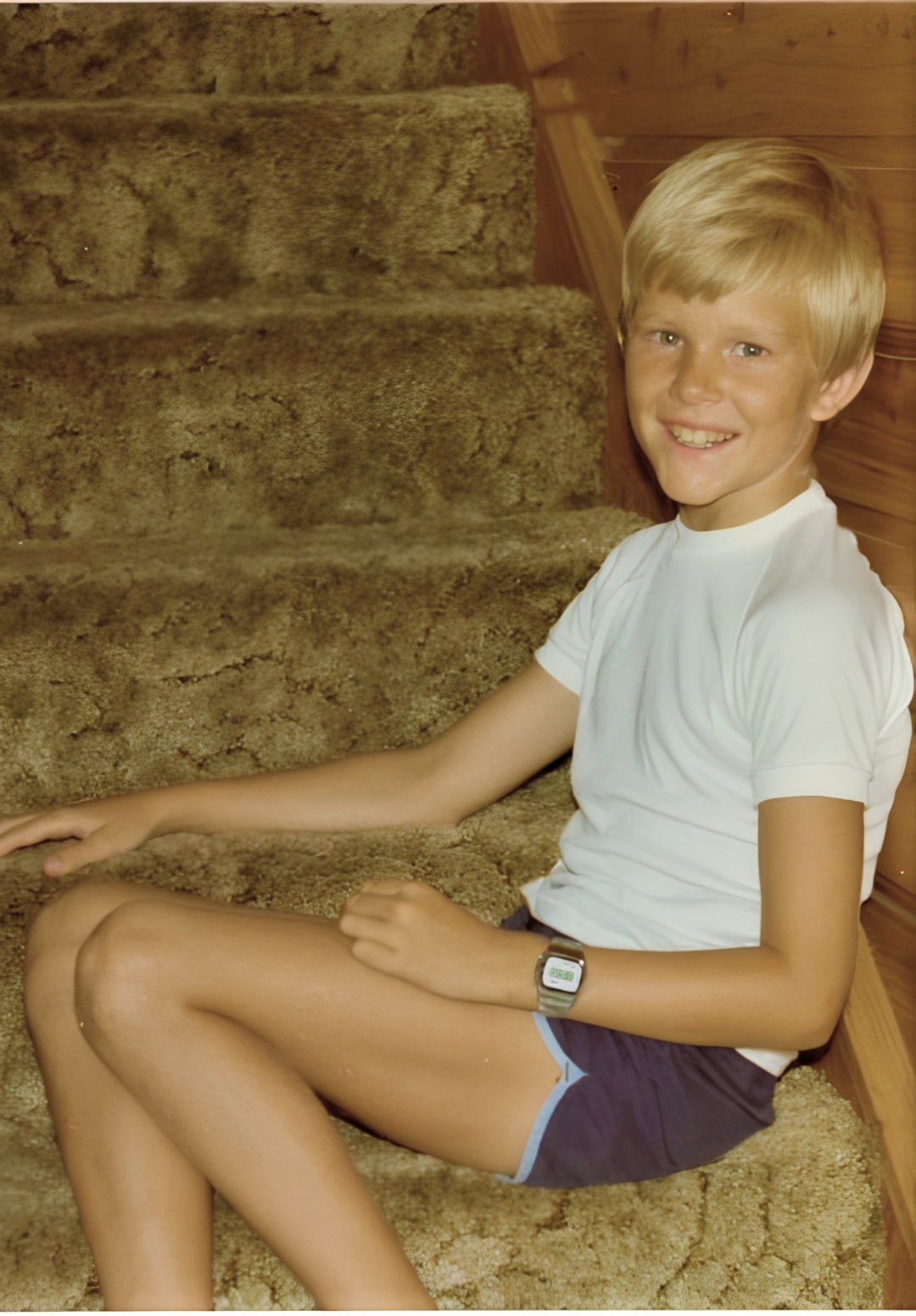 Craig as a boy sitting on carpeted stairs, white t-shirt and navy shorts, wearing a wristwatch
