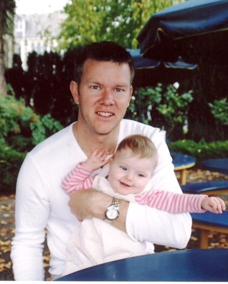 A young Craig holding baby Caitlin, both looking at the camera in a garden
