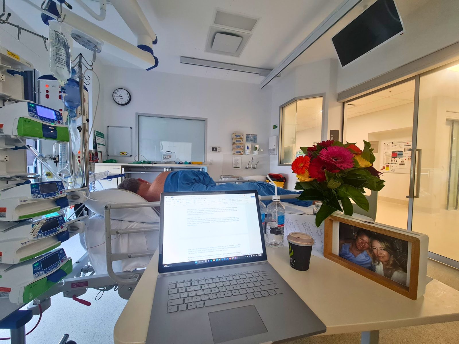 Hospital ICU room with Craig in a bed, a laptop open on the tray table and flowers beside it